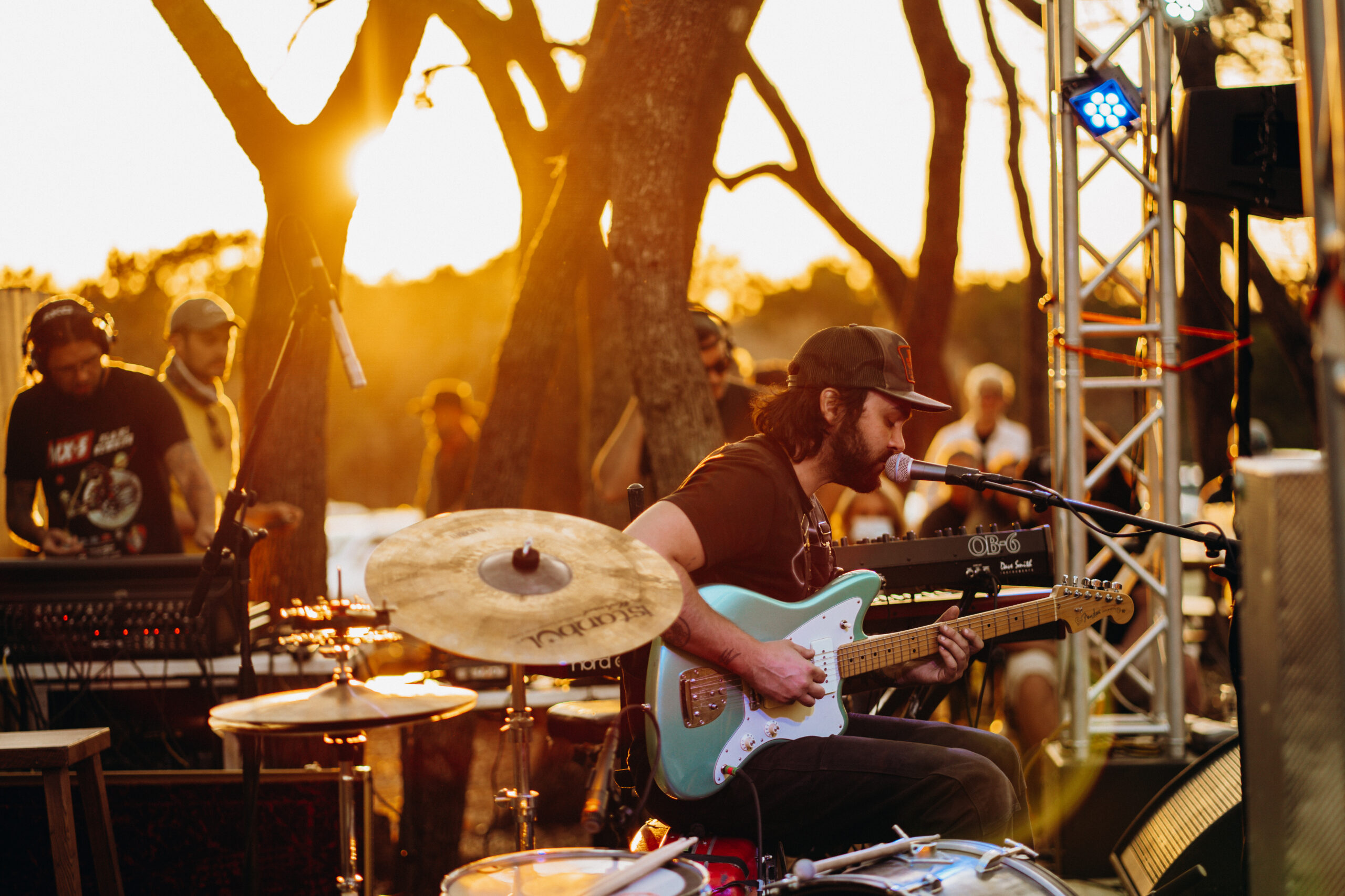 An orange glow from the setting sun in the upper left illuminates a guitar-playing singer at the right, seated next to a drum set and keyboard with a sound technician mixing on the left of beerburg’s outdoor stage in the beer garden; a blurred crowd in the background watches through the oak trees, enjoying the live music.