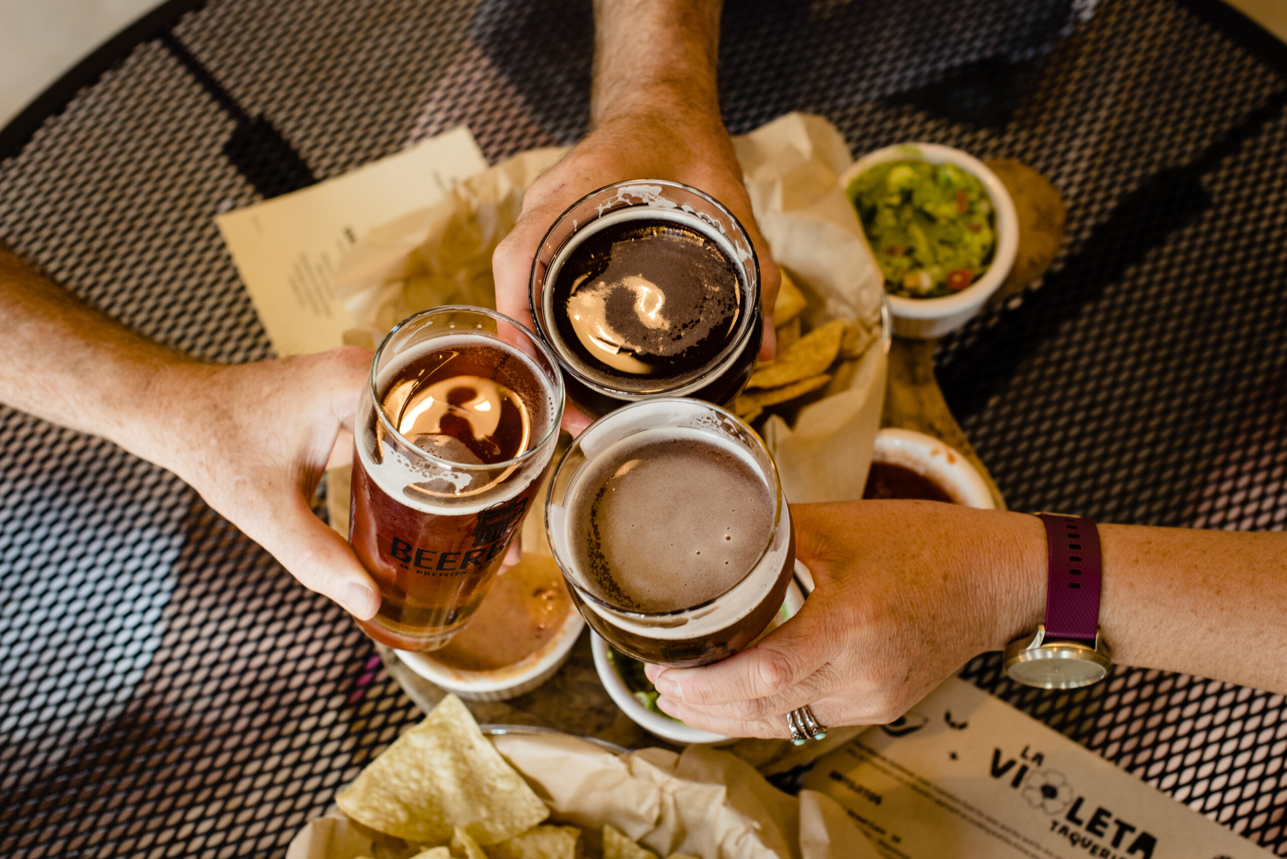 Three hands holding different colored beers in glasses cheers over chips and guacamole; the image promotes shared experience and good times, perfect for corporate team building and celebrations of milestone achievements.