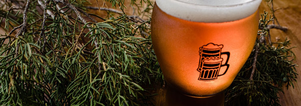 A brightly lit pint of beer, golden in color, with Beerburg’s twin mugs logo in black on the glass, is featured surrounded by juniper branches on a wood tabletop.