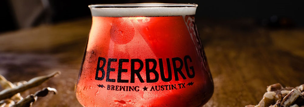 A small, specialty glass holds a bright red beer, highlighted from behind, sitting on a wood table, profiling the unique beers that will accompany this very exclusive reserve tasting package.