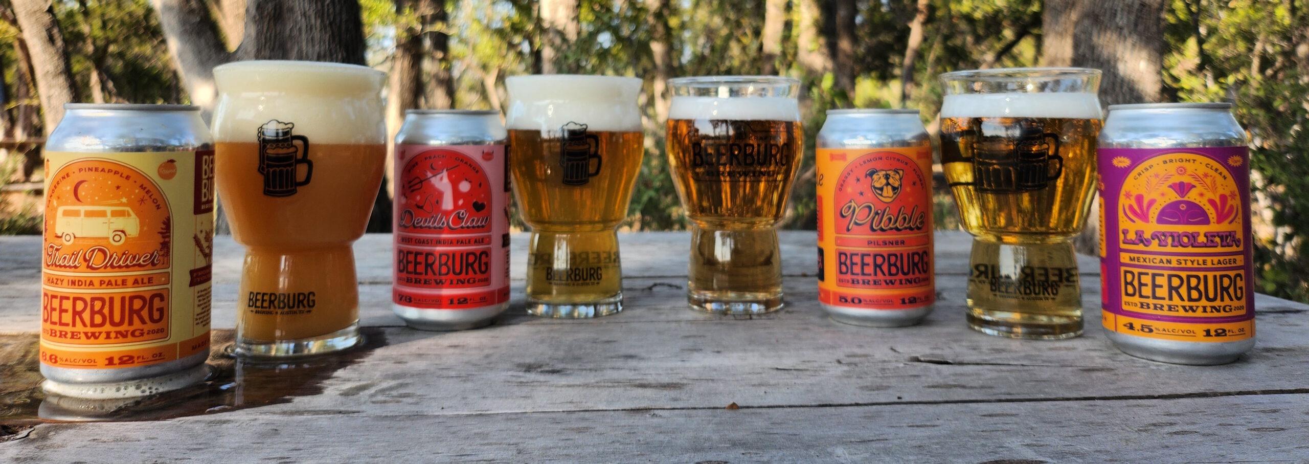 Beerburg’s core 4 beers pictured in cans with brightly colored labels lined up with the beer poured in glasses behind the cans.