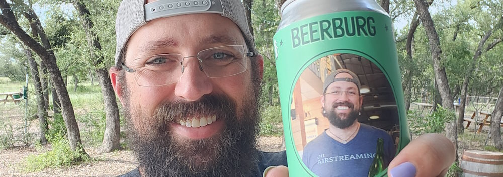 Beerburg owner Ross Nearburg is pictured in a selfie, he holds a 16oz can of beer with a green label depicting his face, showing off the personalization option for custom beer that guests can use for shareable, unique moments during their events, he is standing in front of the beer garden, with tall oaks stretching towards a blue sky behind him.