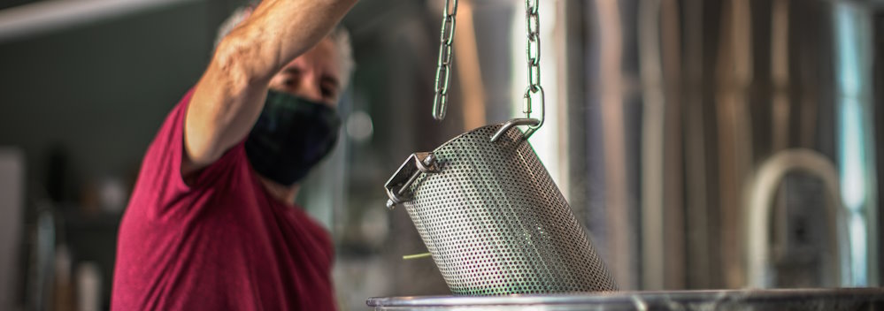 Head Brewer Gino, in a red shirt and wearing a black face mask, lowers a hop strainer into a brew kettle, depicting small batch brewing that would accompany the purchase of the Brew with Us! service.