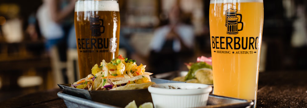 Two beers in branded glasses, one light and one dark, sit behind a metal tray of one of Beerburg’s in-house catering options, tacos, piled high, topped with cotija cheese, and sitting next to a salsa dish, profiling both the food option and style of plating you can expect from Beerburg.